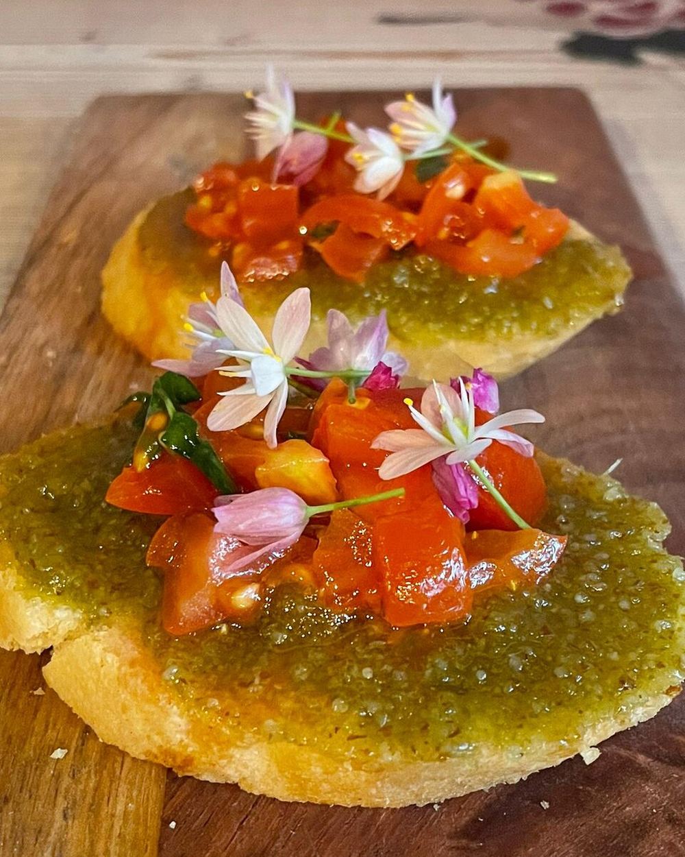 Due crostini su un tagliere di legno, con crema verde, pomodorini datterini a pezzetti e fiori commestibili bianchi e rosa.