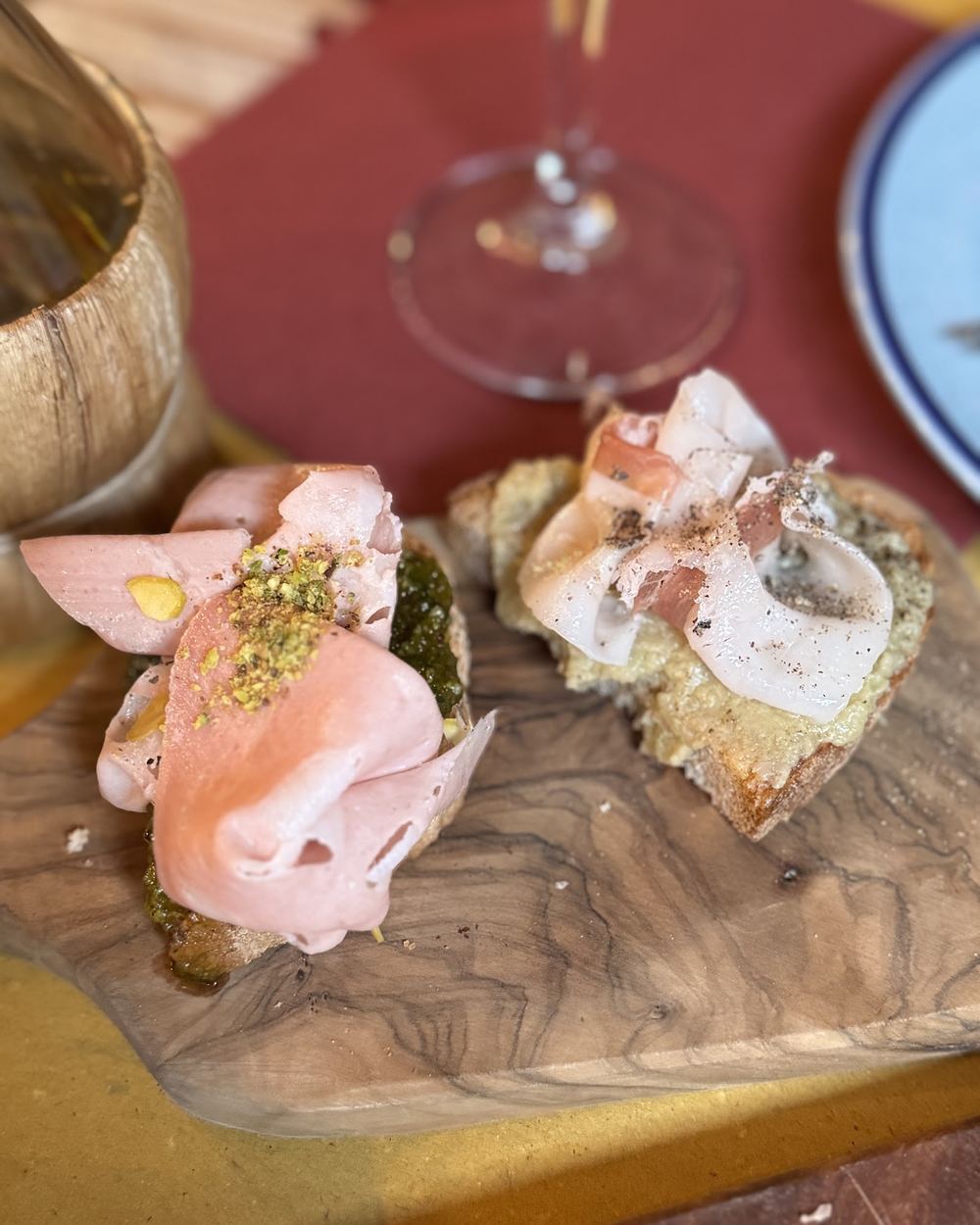 Due crostini su un tagliere di legno, uno con crudo e crema chiara, l'altro con salsa scura e granella verde.