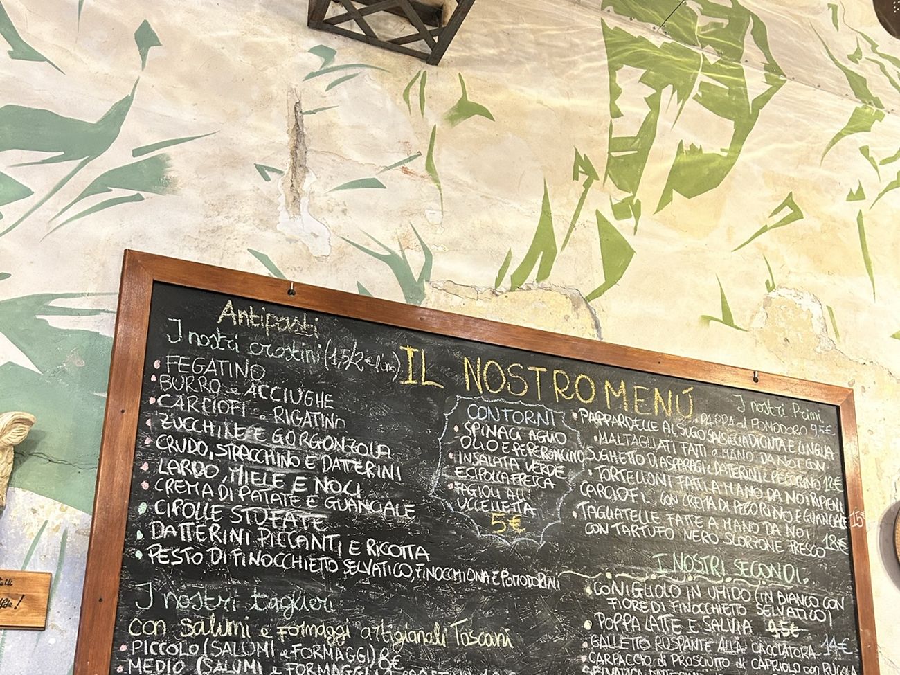 La lavagna del locale, intitolata Il nostro men&ugrave;, scritta a gesso bianco e giallo e appesa a una parete di calce con foglie verdi dipinte.