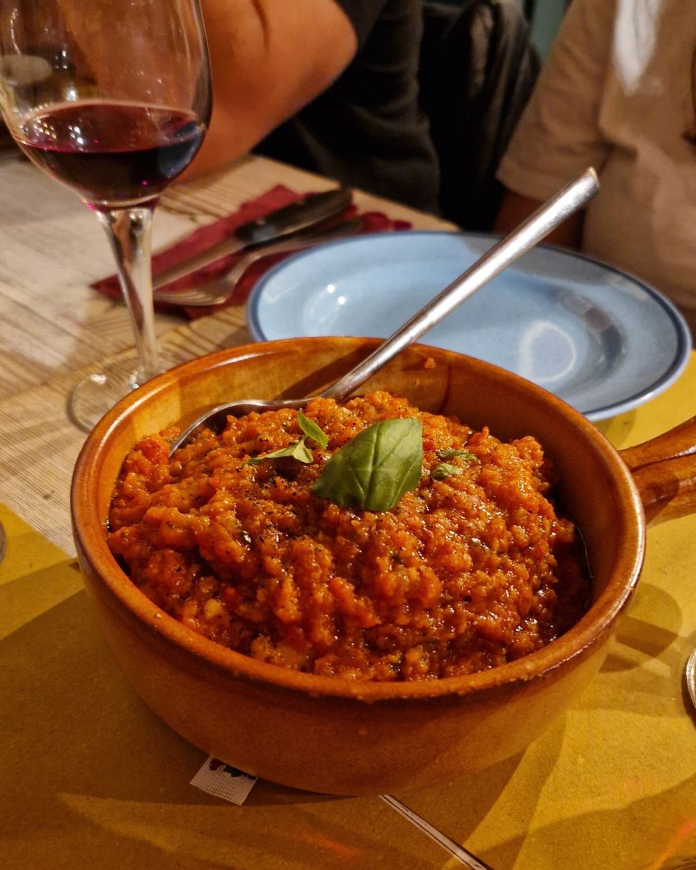 Una cocotte di terracotta piena di pappa al pomodoro con una foglia di basilico sopra, accanto a un calice di vino rosso.