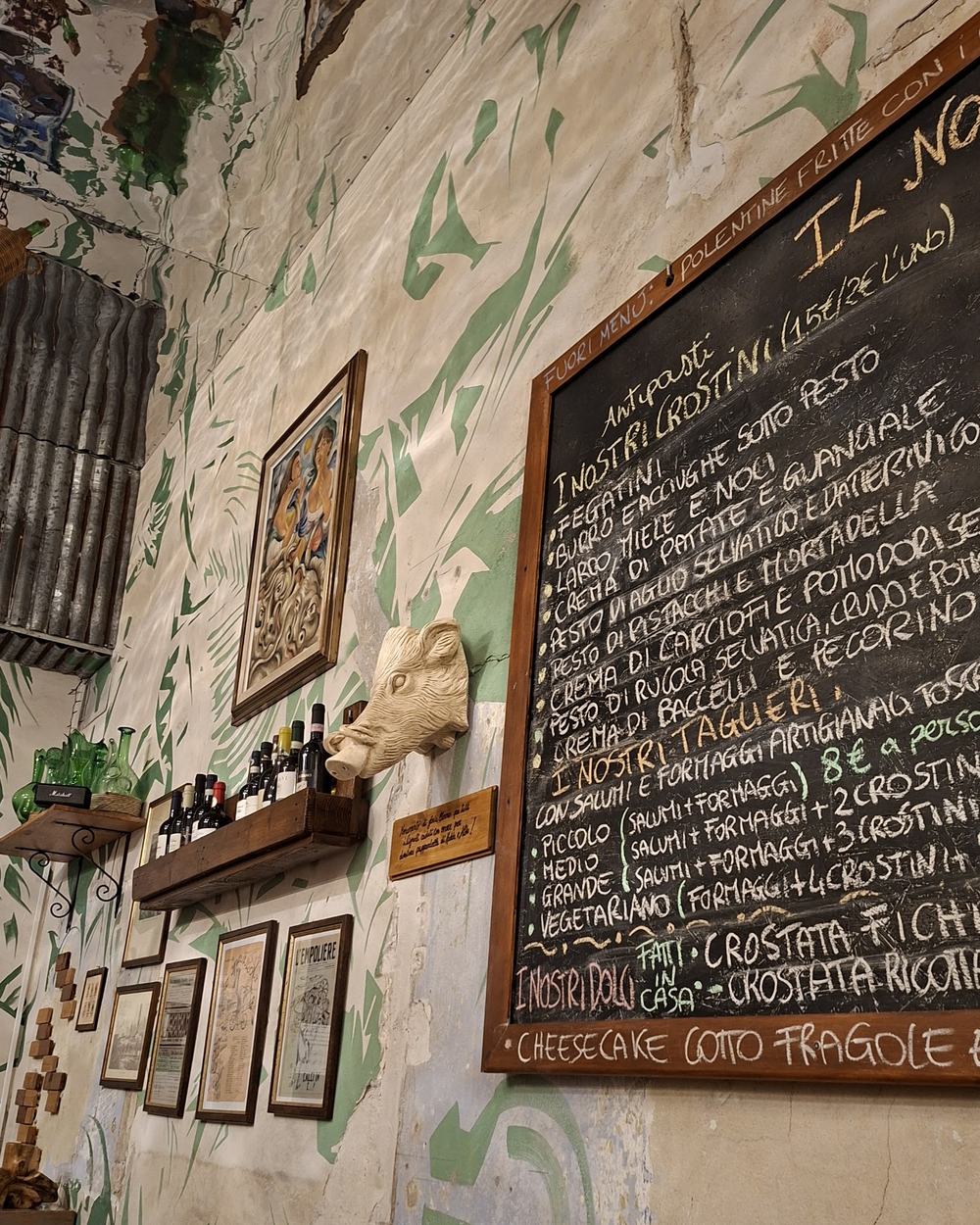 La parete della sala: calce chiara con foglie verdi dipinte, stampe incorniciate, una mensola di distillati e il bordo della lavagna del menu.