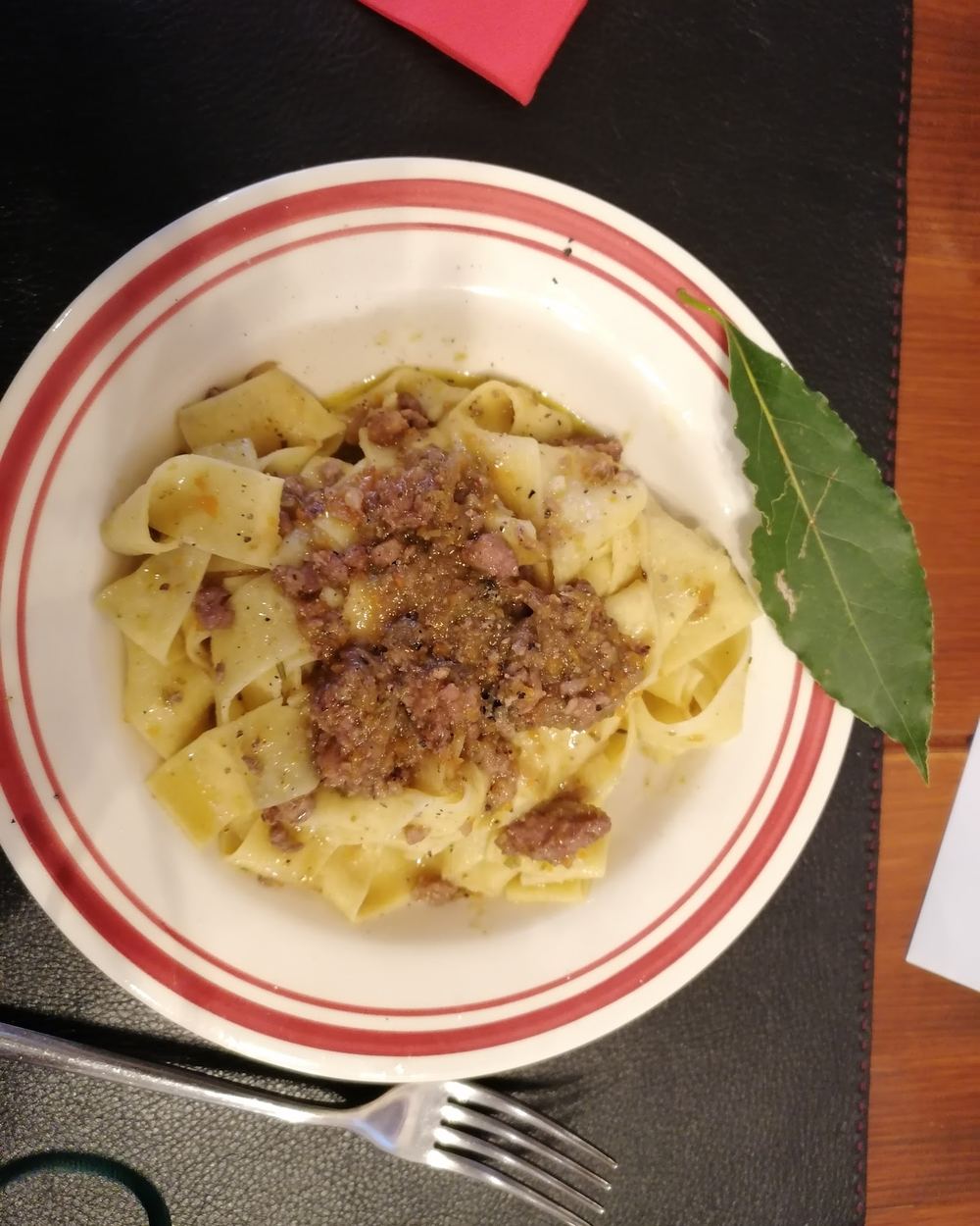 Pappardelle in un piatto bianco dal bordo rosso, condite con un rag&ugrave; di carne e una foglia di alloro.