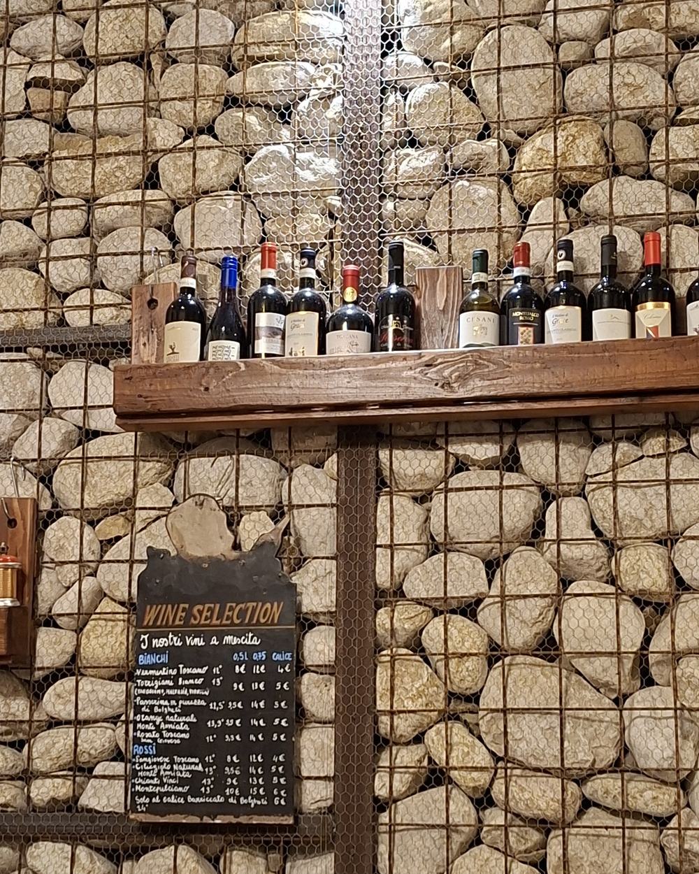 Una mensola di legno con bottiglie di vino toscano fissata a una parete di sassi in gabbioni, e sotto una lavagnetta intitolata Wine Selection con i vini al calice.